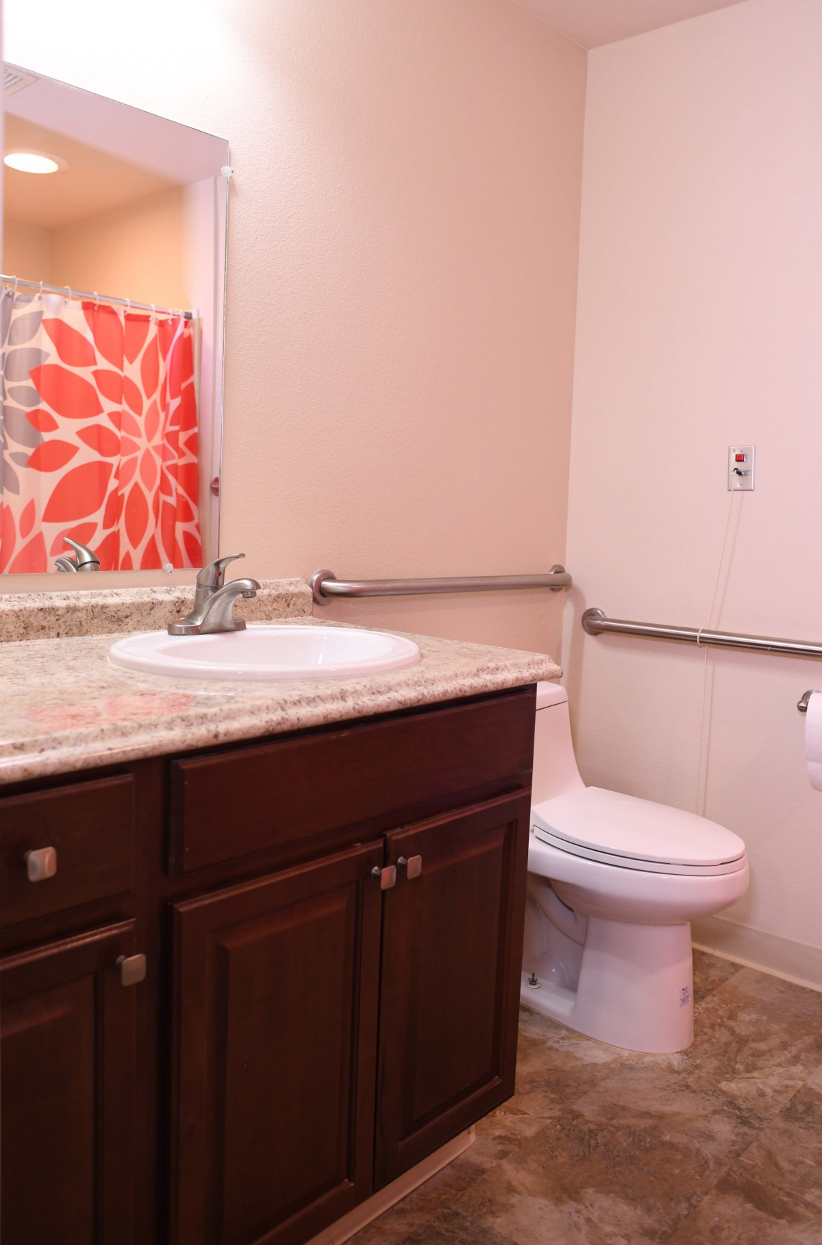 A bathroom with a wooden vanity, sink, and toilet, featuring a red floral shower curtain and safety handrails.