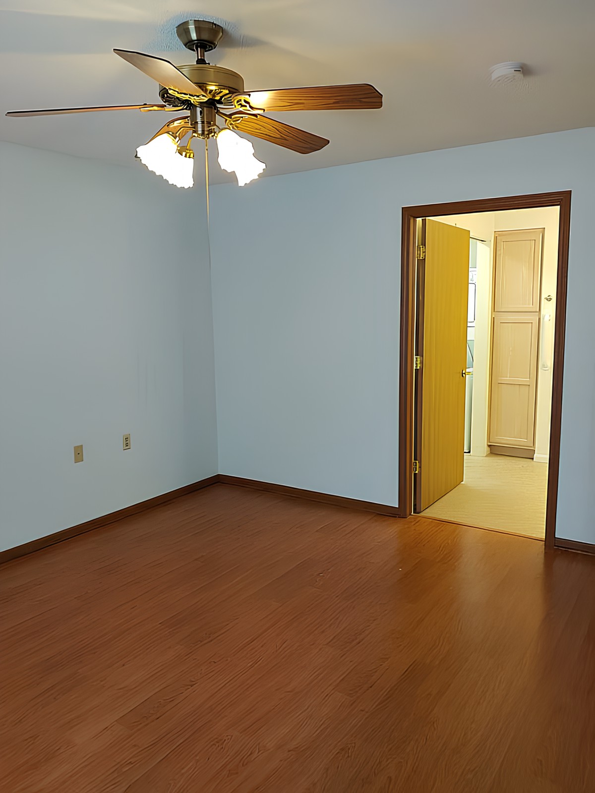 An empty room with light blue walls, wood flooring, and a ceiling fan, leading to an adjoining bathroom.