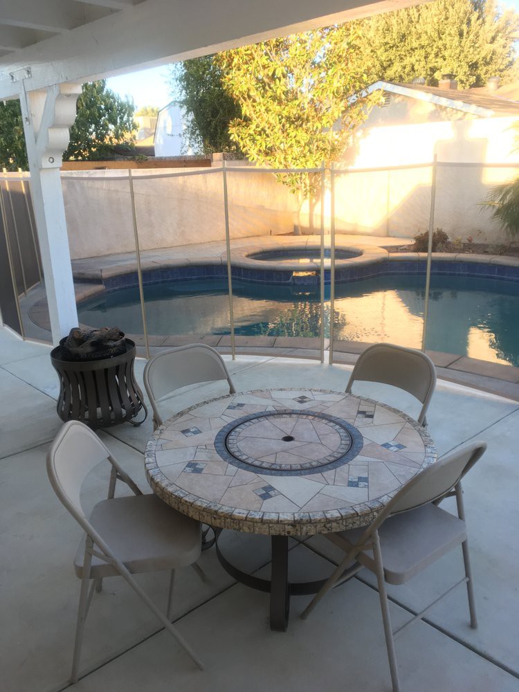 A backyard patio with a round table, chairs, and a view of the swimming pool enclosed by a safety fence.