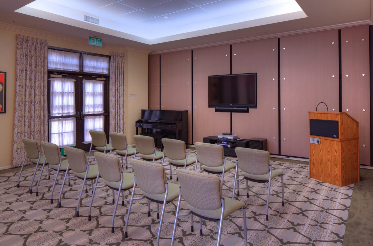 A conference room setup with rows of chairs facing a television and a podium, featuring a piano in the corner.