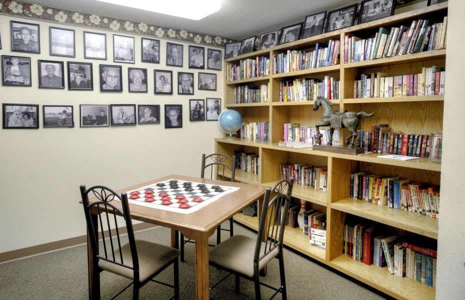 A cozy library room with bookshelves, a checkers board on the table, and framed photos on the wall.