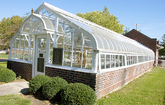 A brick-based greenhouse with glass panels, surrounded by greenery.