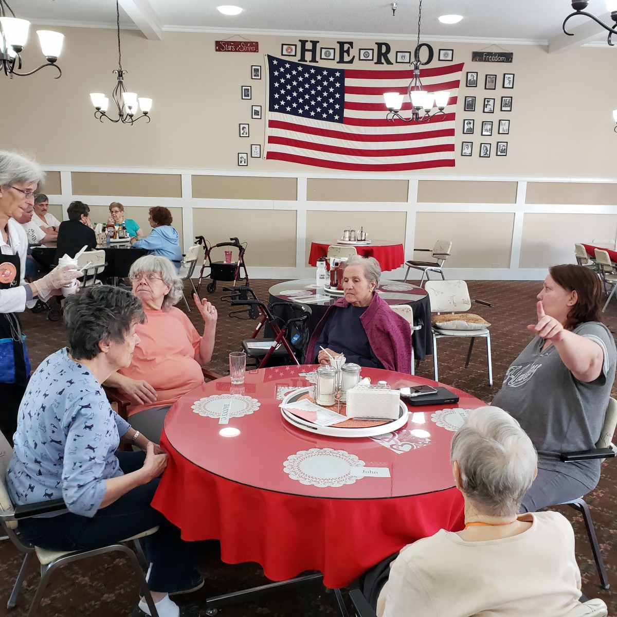 A group of seniors sitting around a red tablecloth, engaged in conversation with an American flag displayed in the background.
