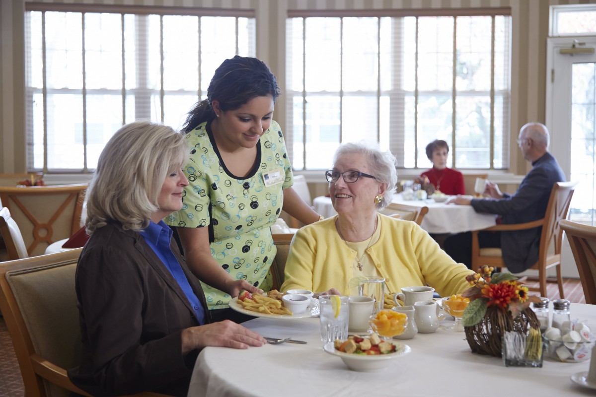 A caregiver interacting with two elderly women in a nursing home dining room.