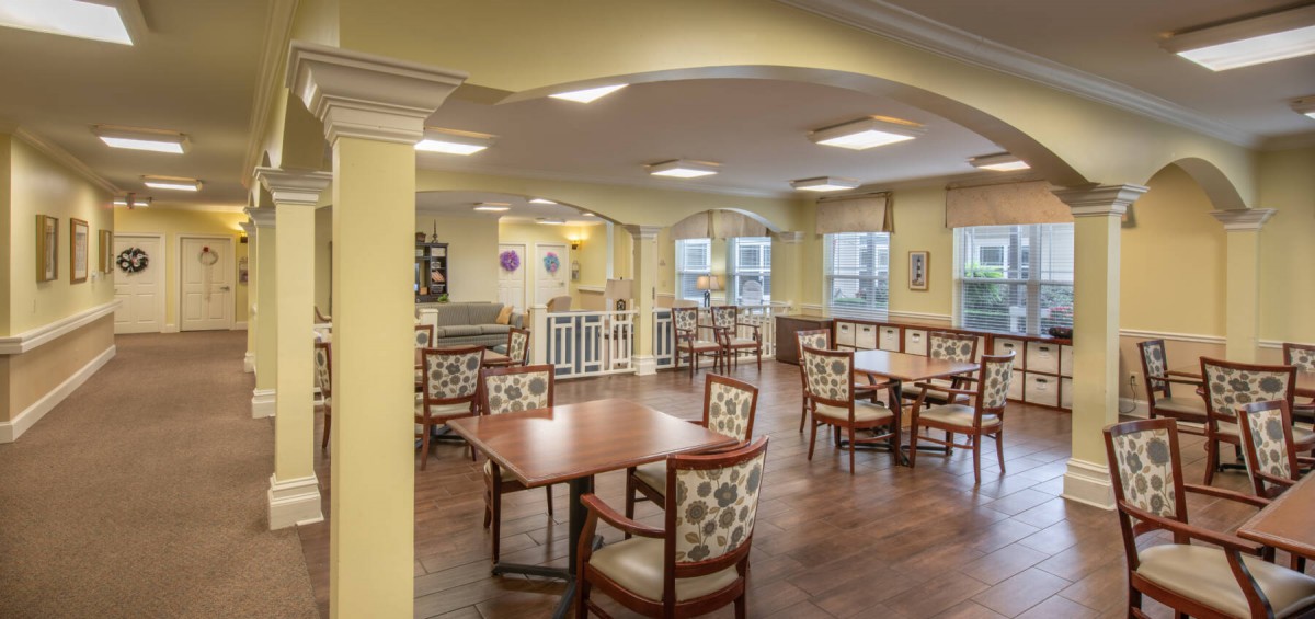 A well-lit dining area with multiple tables and chairs, featuring large windows and wooden flooring.