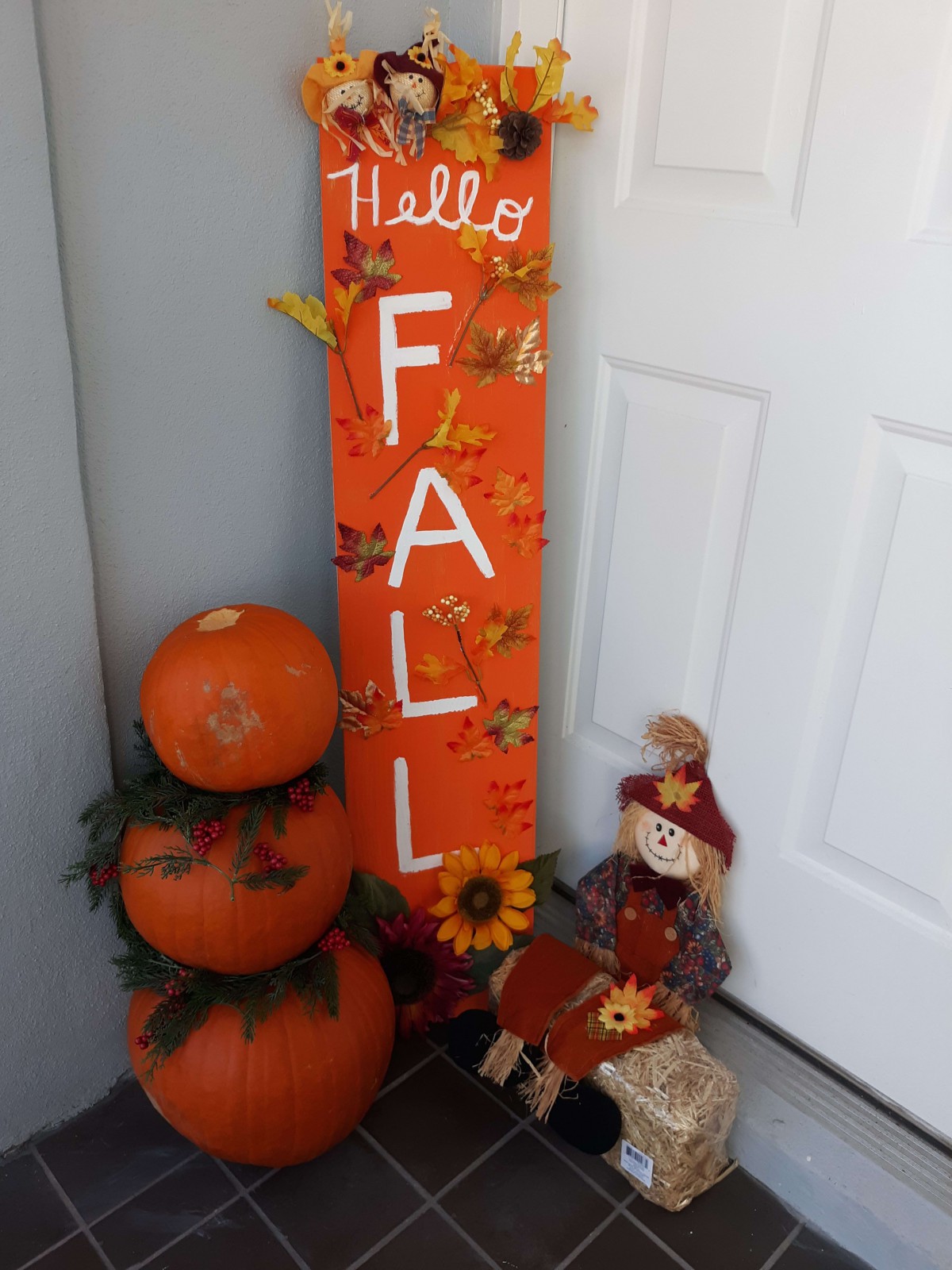 A welcoming fall display with stacked pumpkins, a colorful 'Hello Fall' sign, and a small scarecrow.