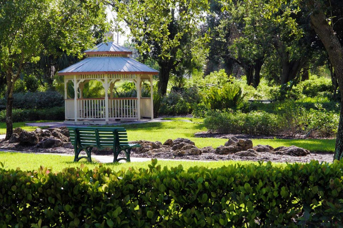 A serene park scene with a gazebo and a green bench surrounded by lush greenery.