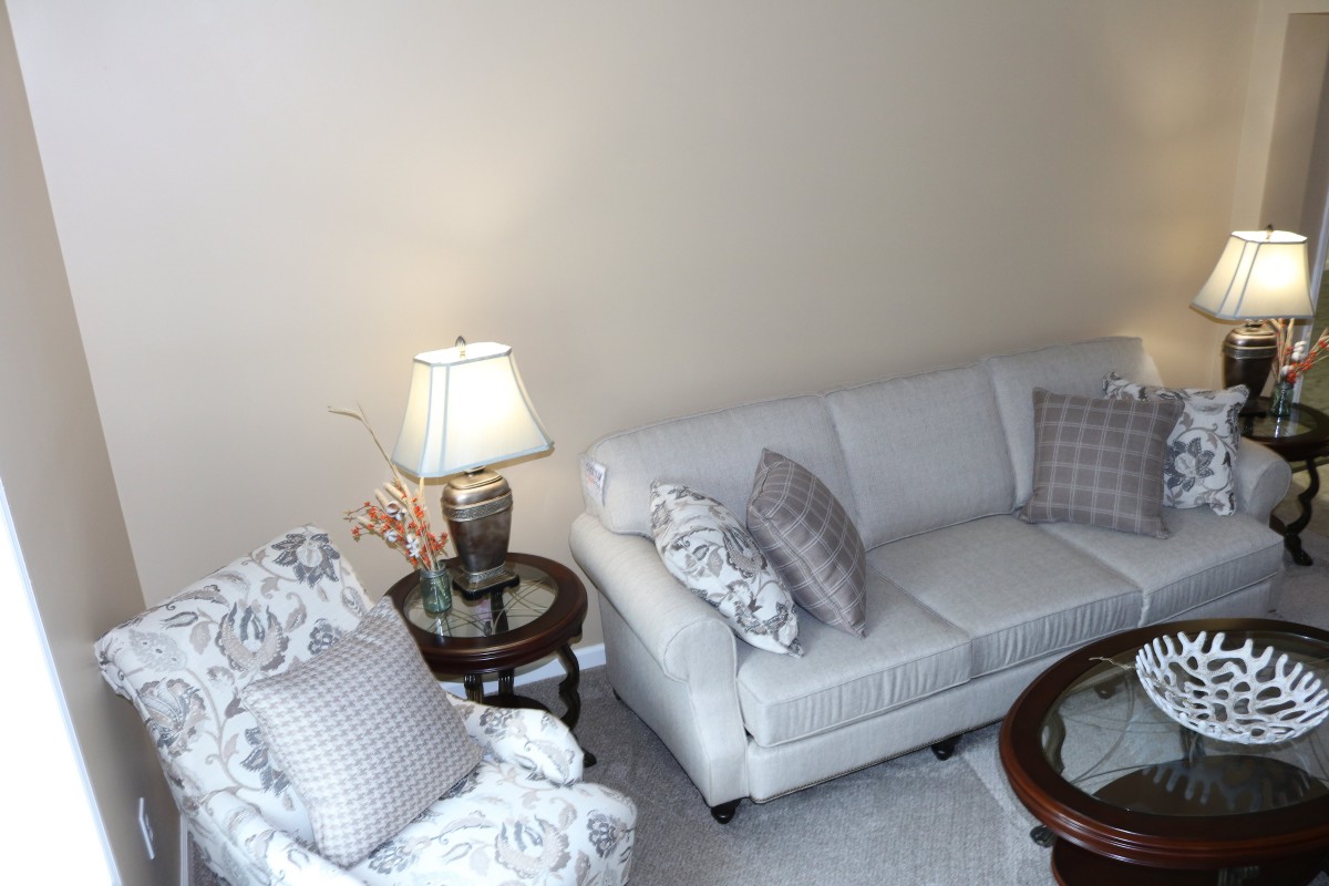 A cozy living room with a white sofa and matching armchair, adorned with patterned cushions and accompanied by a side table with a lamp.
