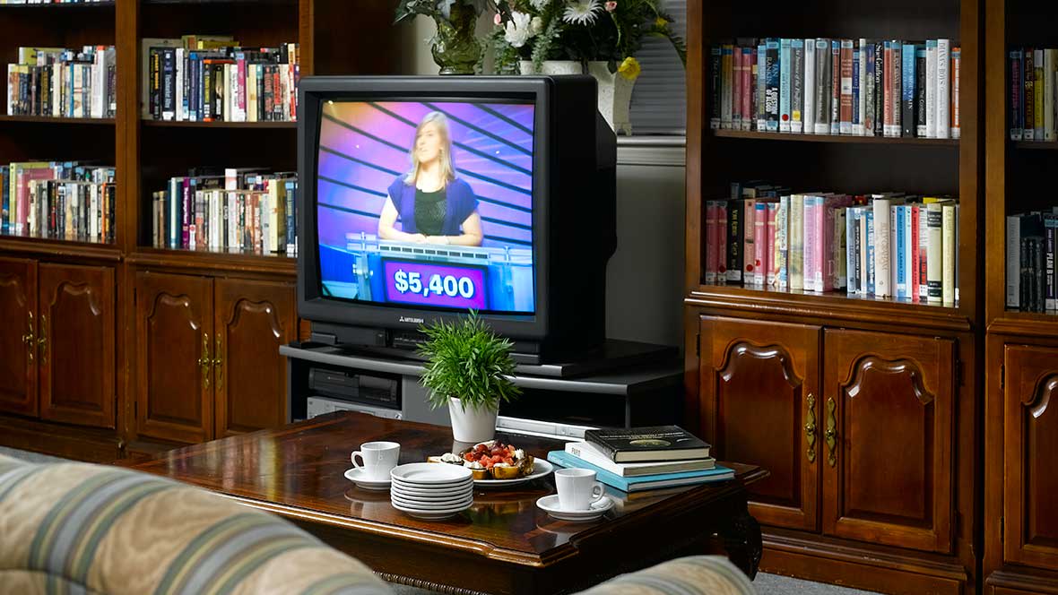 A cozy living room with a television showing a game show, surrounded by bookshelves and a coffee table with cups and snacks.