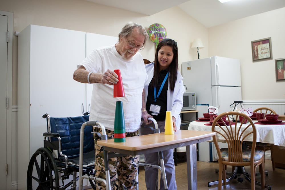 An elderly man receiving physical therapy assistance, stacking cups with guidance from a healthcare provider.