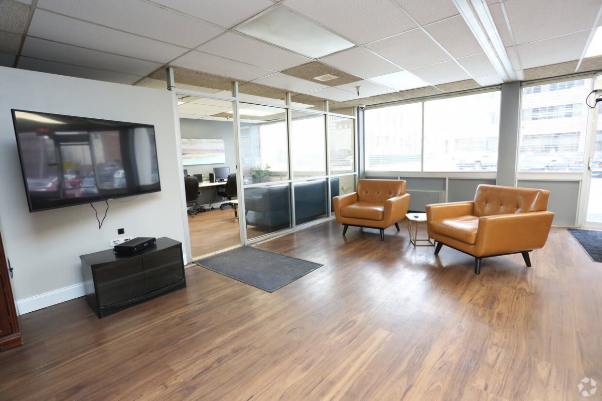 A modern office waiting area with glass walls, two leather chairs, a television, and wooden flooring.