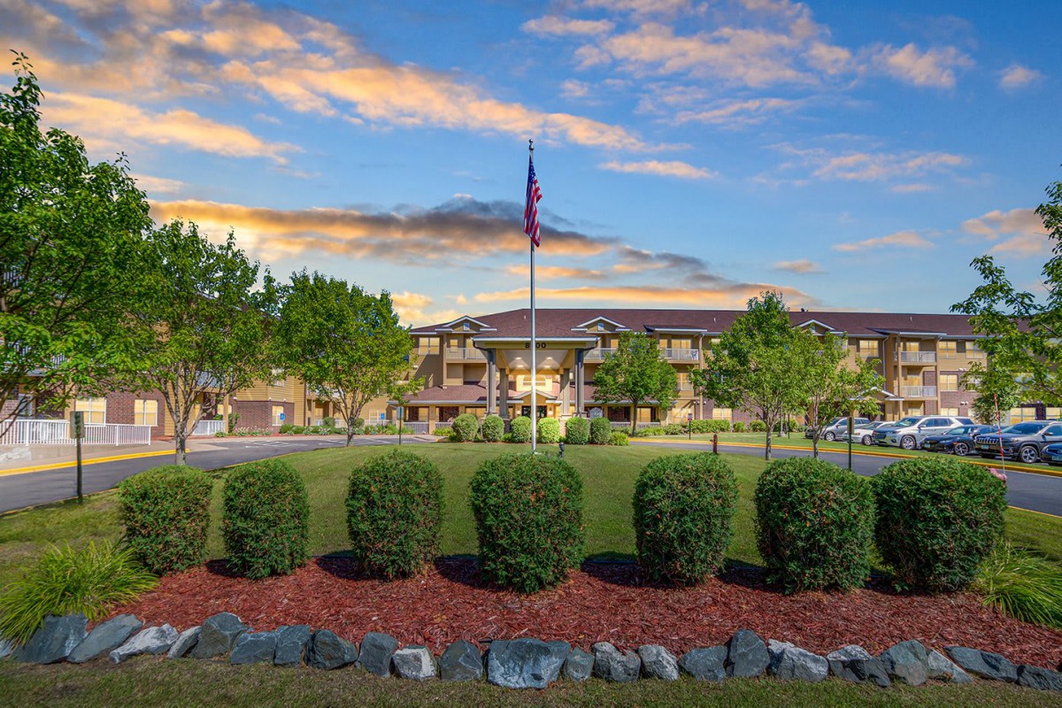 A scenic view of a building with a flagpole, surrounded by landscaped greenery and set against a backdrop of colorful clouds at sunset.