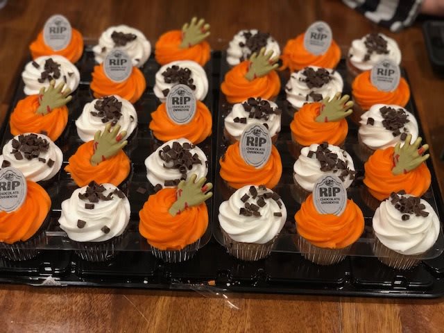 A tray of Halloween-themed cupcakes decorated with orange and white frosting, tombstone plaques, chocolate chips, and zombie hands.