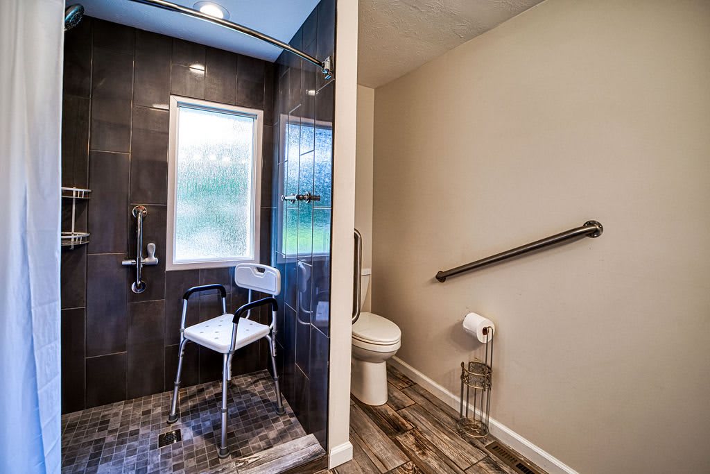 A modern bathroom featuring an accessible shower with a chair and a toilet equipped with a grab bar.