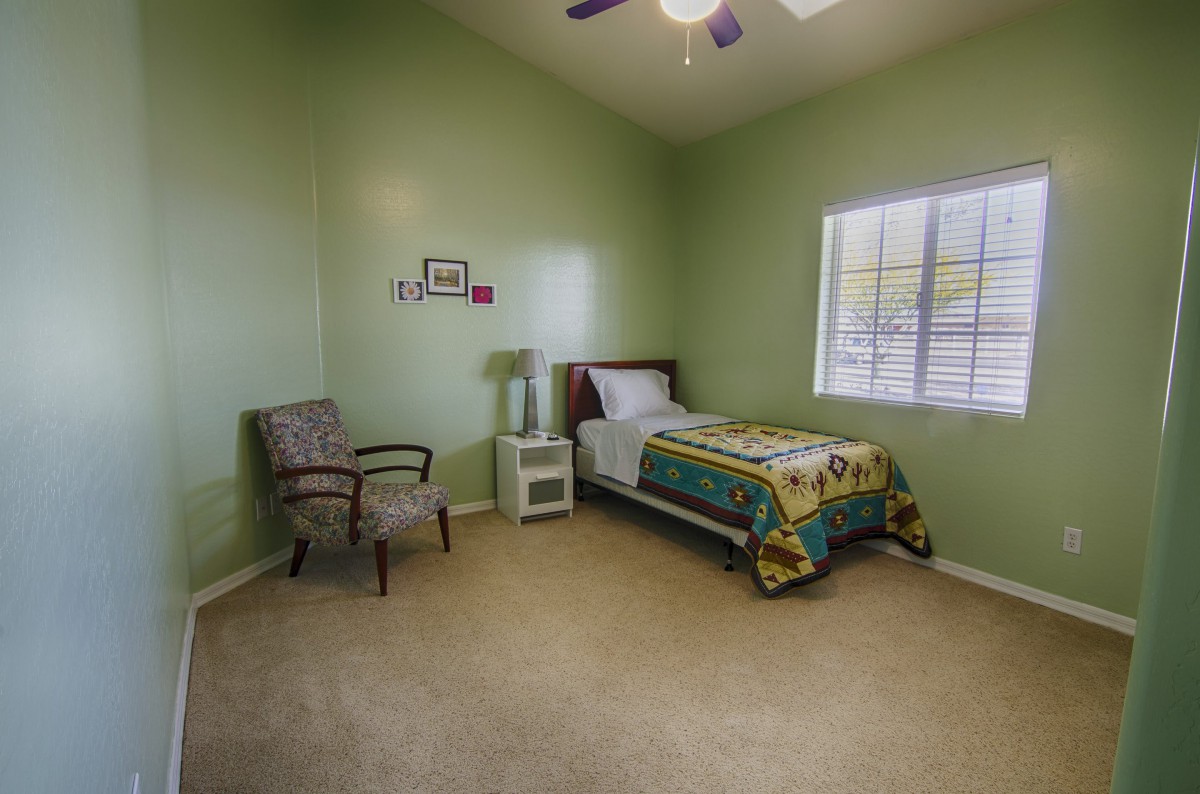 A minimalist bedroom with green walls and a single bed, featuring a chair and a lamp by the window.
