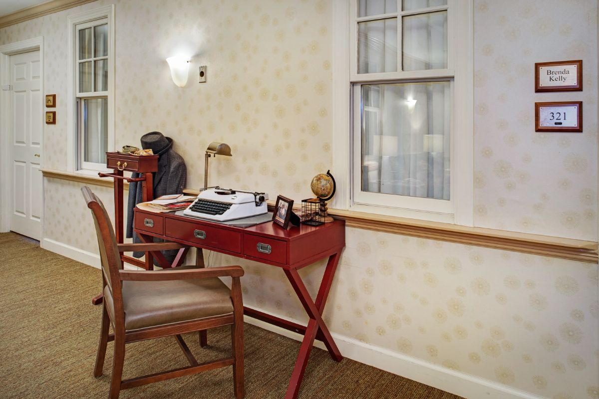 A vintage office setup with a typewriter on a red desk, accompanied by a wooden chair and a nearby bookshelf.