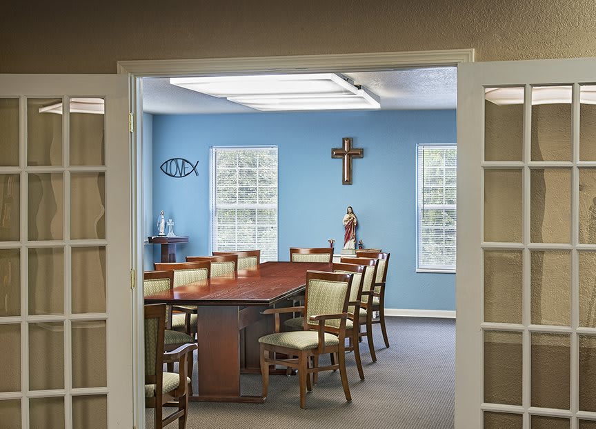A spacious meeting room with a long wooden table and several chairs, featuring a blue wall decorated with a cross and large windows.