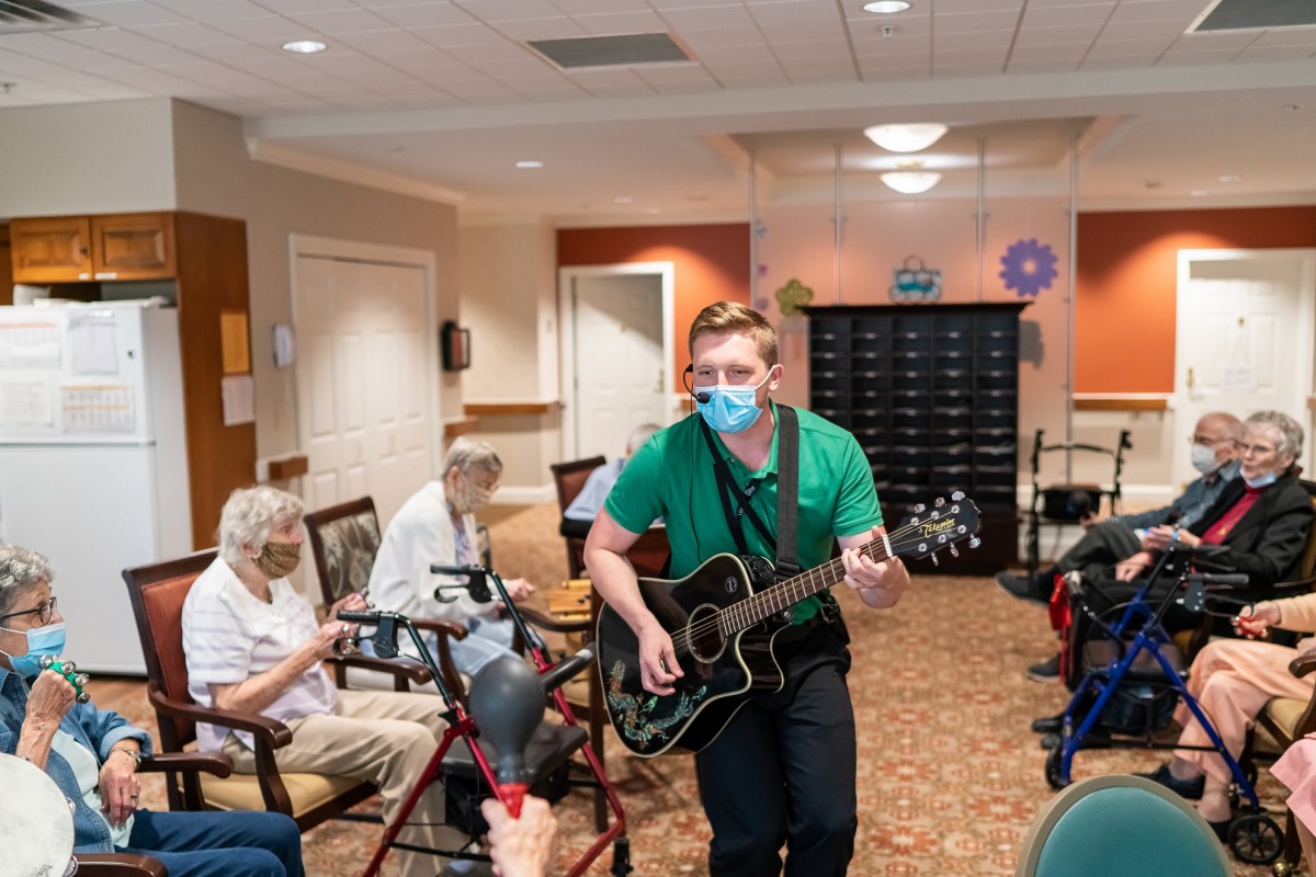 A musician performs for residents at a nursing home, playing guitar and wearing a mask.