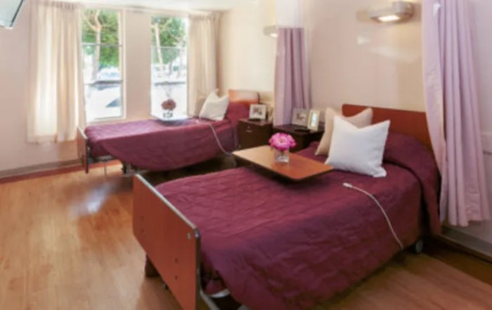 A hospital room with two neatly arranged beds, featuring purple bedspreads and a window view.