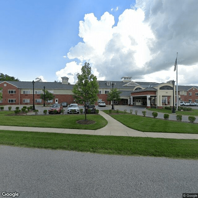 A view of a large brick building with a parking lot in front, surrounded by green lawns and a pathway under a cloudy sky.