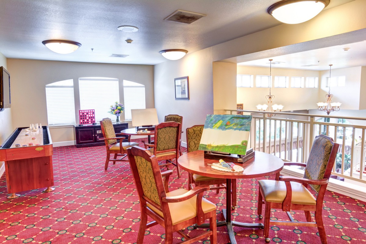 A cozy lounge area with tables and chairs, carpeted flooring, and ample natural light from large windows.
