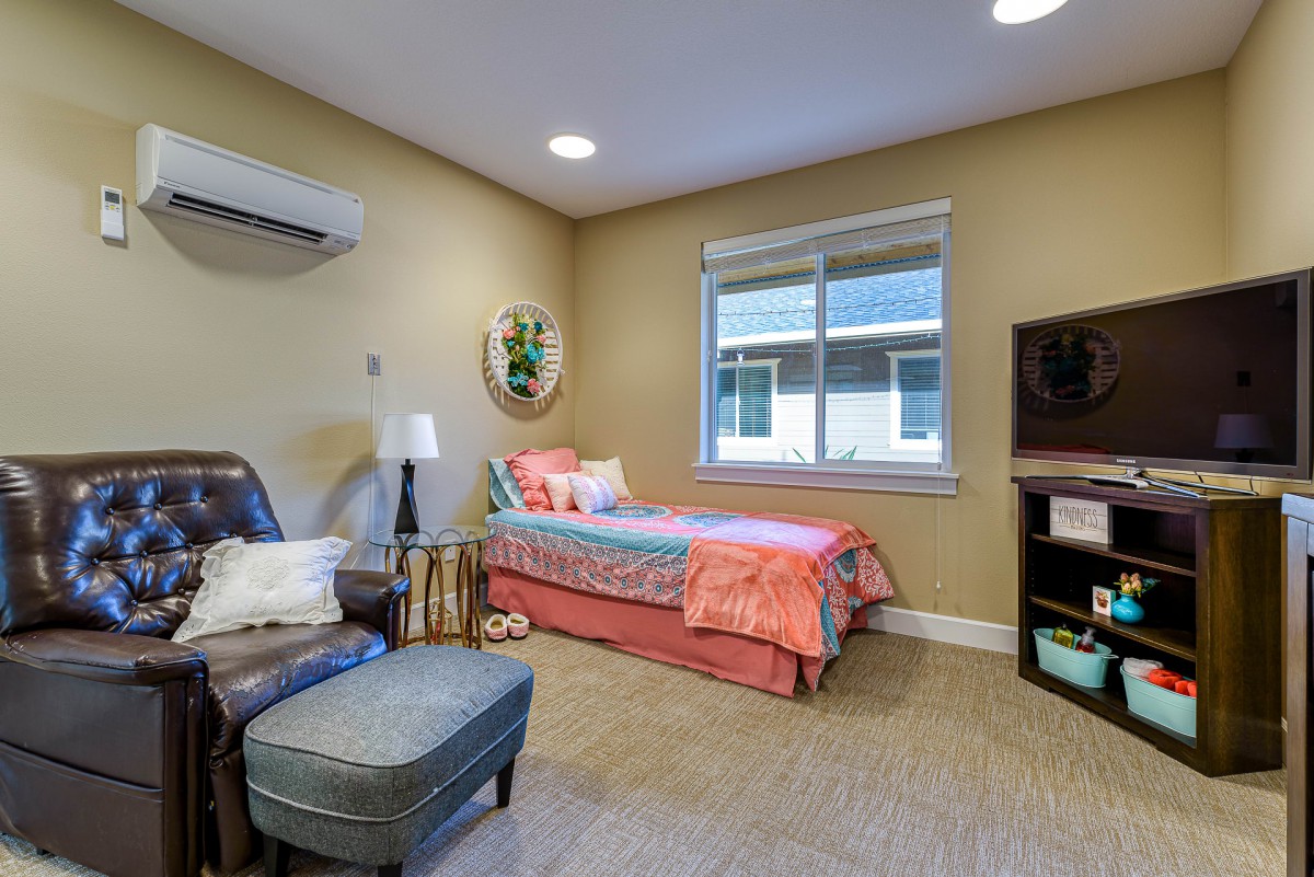 A cozy bedroom with a single bed, armchair, and bookshelf, featuring an air conditioner and a window letting in natural light.
