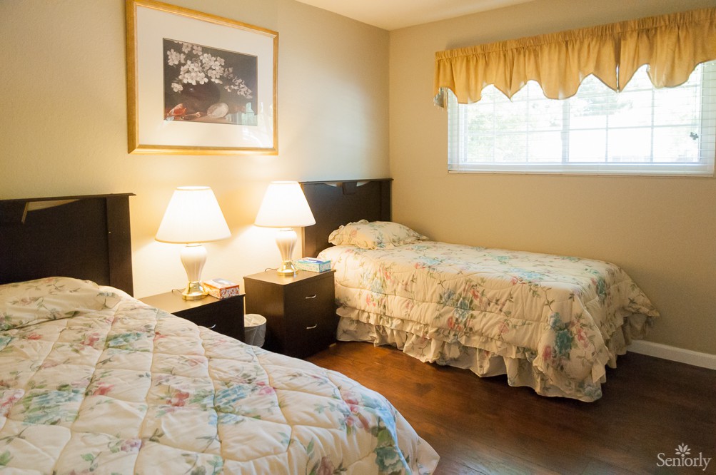 A cozy bedroom with two twin beds, each adorned with floral bedspreads. Two lamps on a nightstand illuminate the room, which features a window with yellow curtains and framed artwork on the wall.