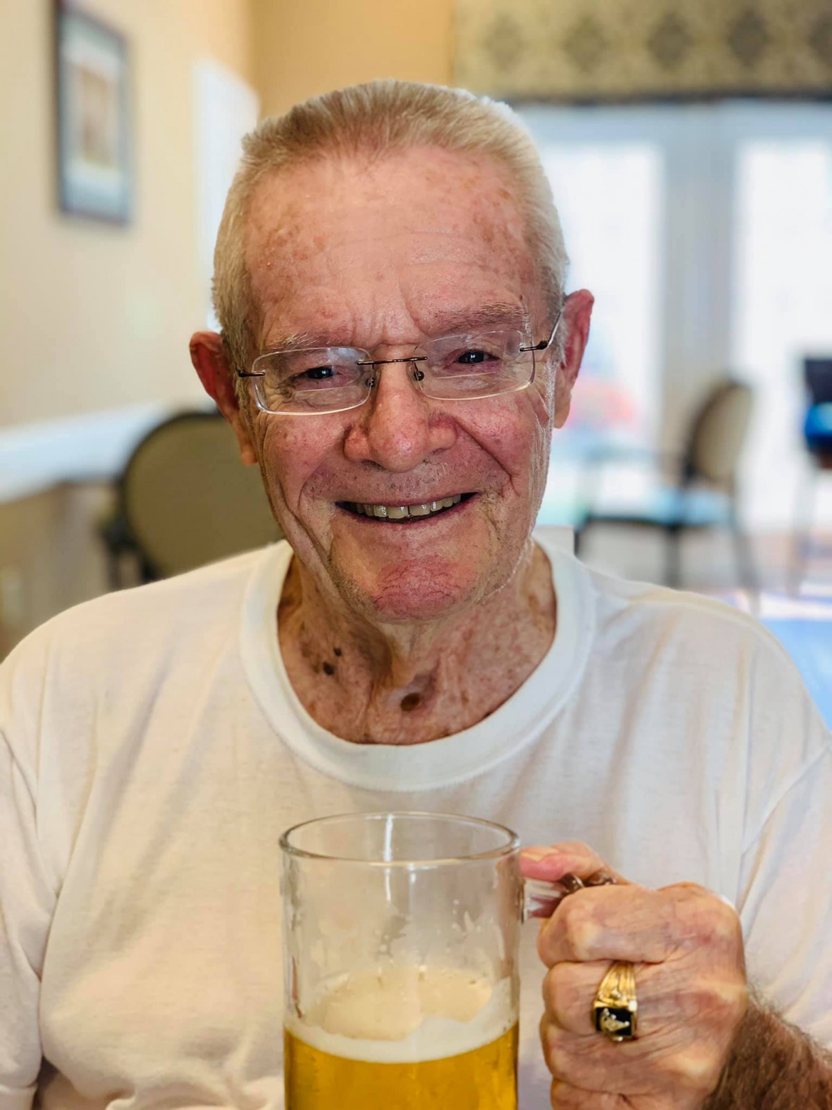 An elderly man wearing glasses is smiling and holding a cup indoors.