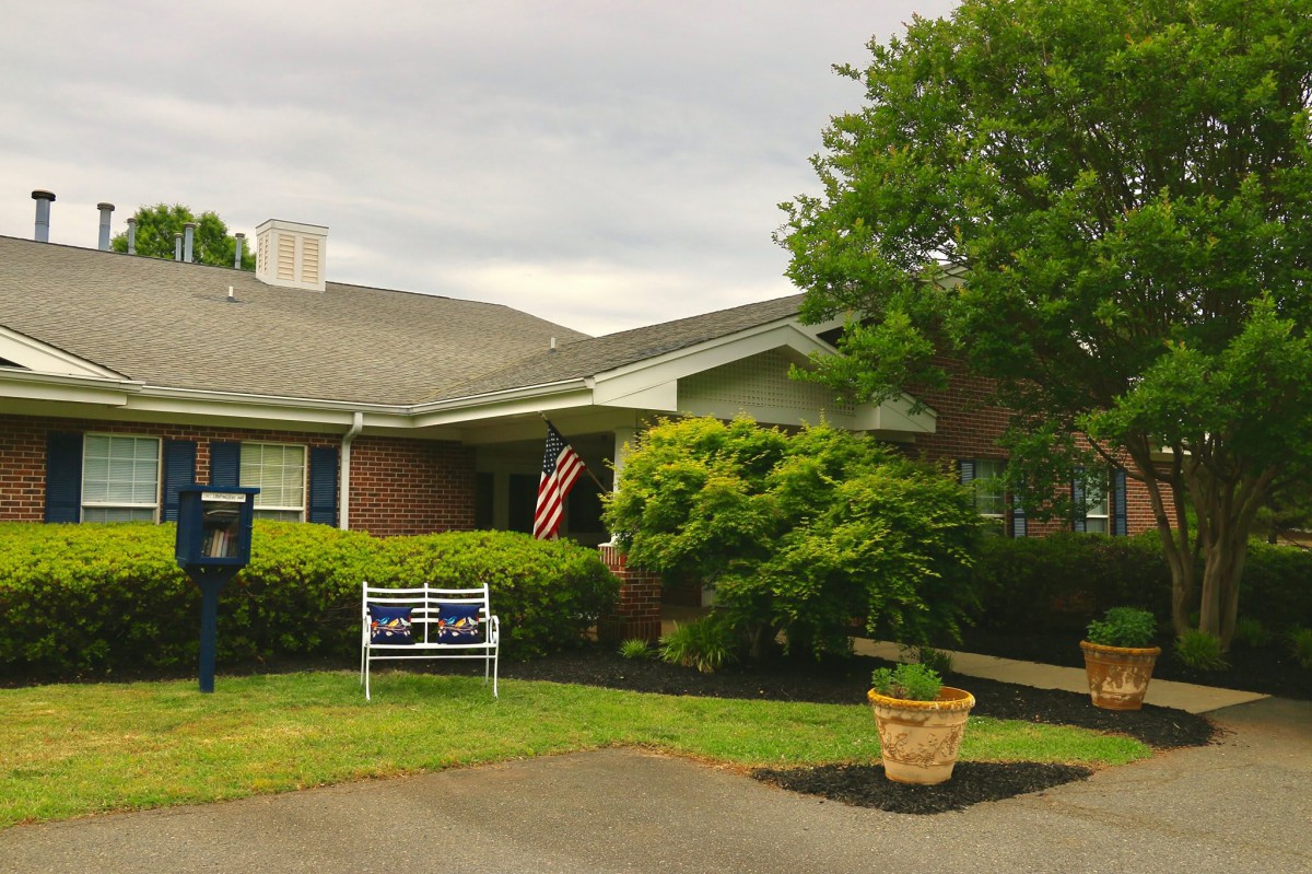 A quaint residential house with a garden, displaying an American flag and a bench on the front lawn.