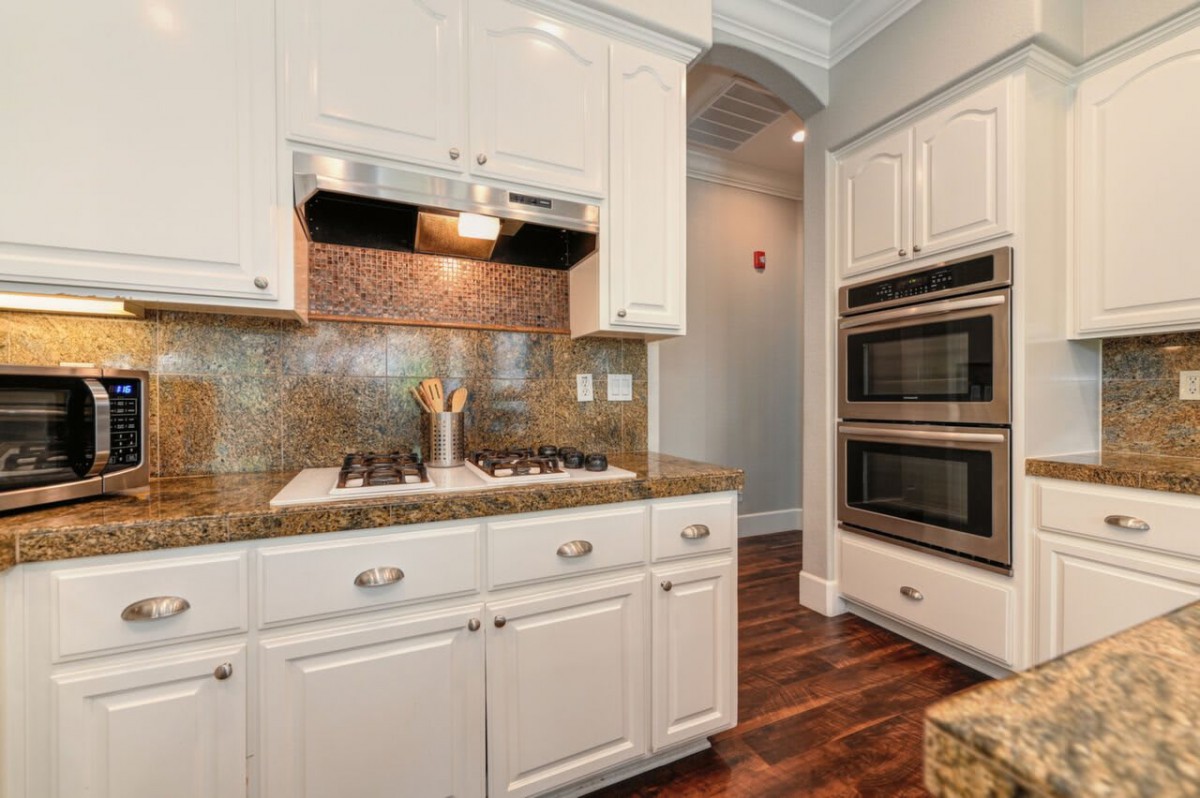 A modern kitchen with white cabinets, double ovens, and a stovetop on a granite countertop.