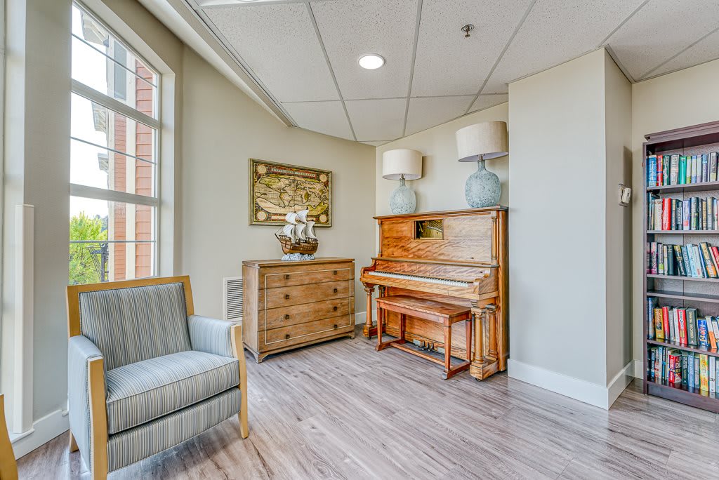 A cozy room interior featuring a wooden piano, chest of drawers, and comfortable chair by the window.