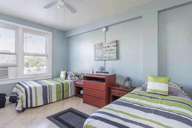 A modern bedroom featuring two twin beds with striped bedding and a central dresser, complete with wall artwork and natural light from a large window.