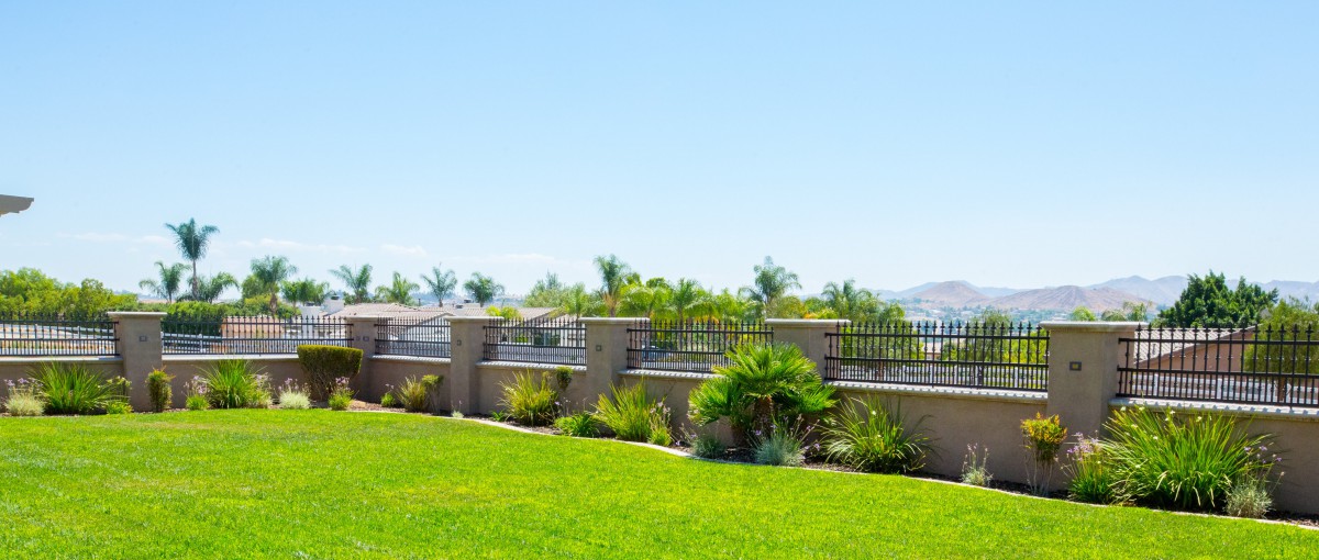 A scenic view of a well-maintained lawn with lush plants and a decorative fence under a clear blue sky.