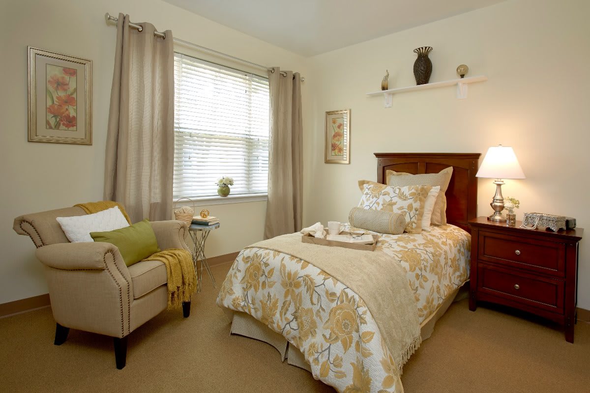 A cozy bedroom with a single bed, patterned bedding, an armchair by the window, and warm lighting.