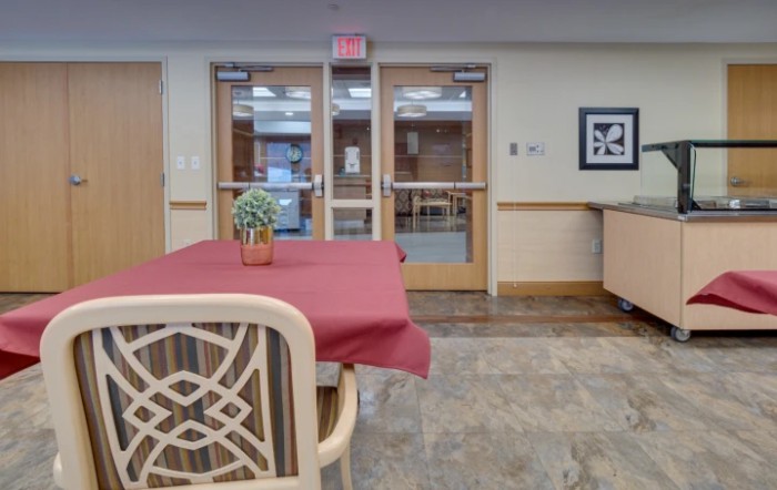 A cozy interior space with a table and chair facing wooden double doors, adorned with an exit sign and small plant centerpiece.