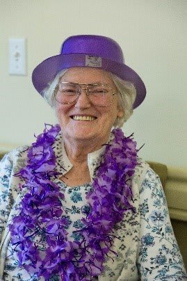 An elderly woman wearing a purple hat and lei necklace smiles warmly.