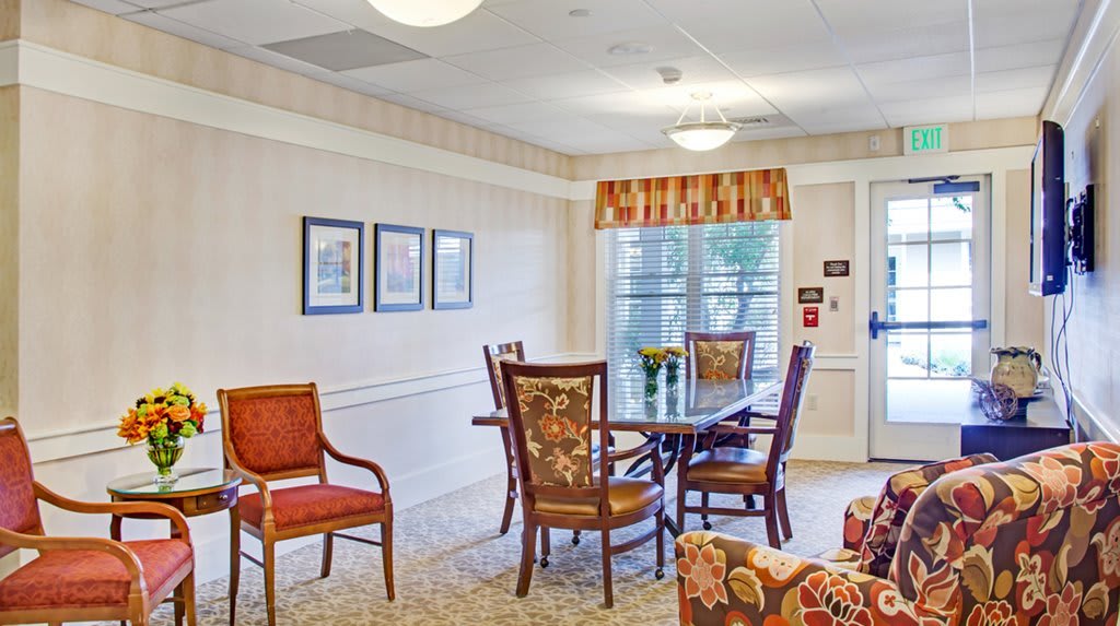 A cozy living room setup with a table surrounded by chairs, wall decorations, and large windows.