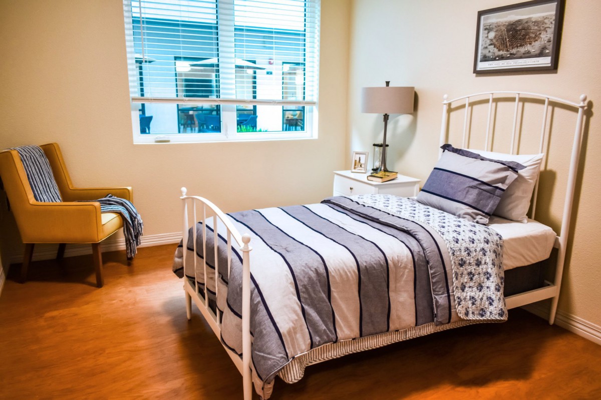 A cozy bedroom with a neatly made bed, a bedside table with a lamp, and a window providing natural light.