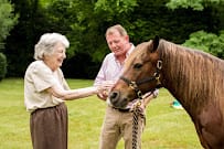 An elderly woman and a man interacting with a horse outdoors.