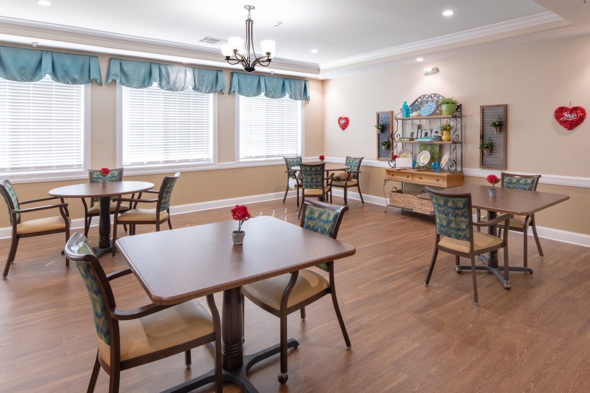 A cozy dining room with neatly arranged tables and chairs, featuring large windows with curtains and a chandelier.