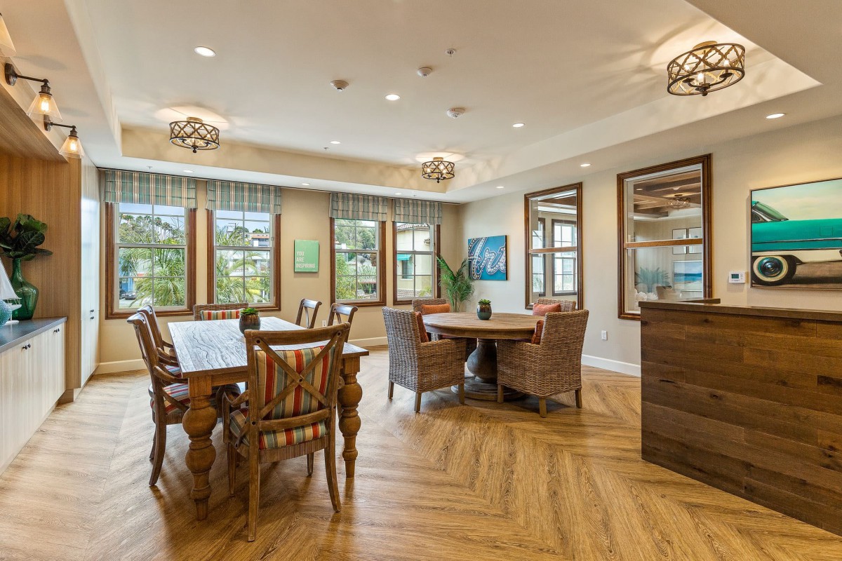 A well-lit dining area with wooden flooring, featuring tables and chairs next to large windows.