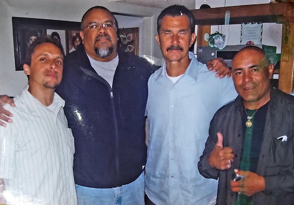 A group of four men standing together indoors, dressed in casual clothing.