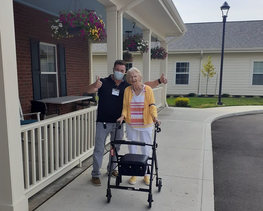 A caregiver assists an elderly person using a walker outside a residential building.