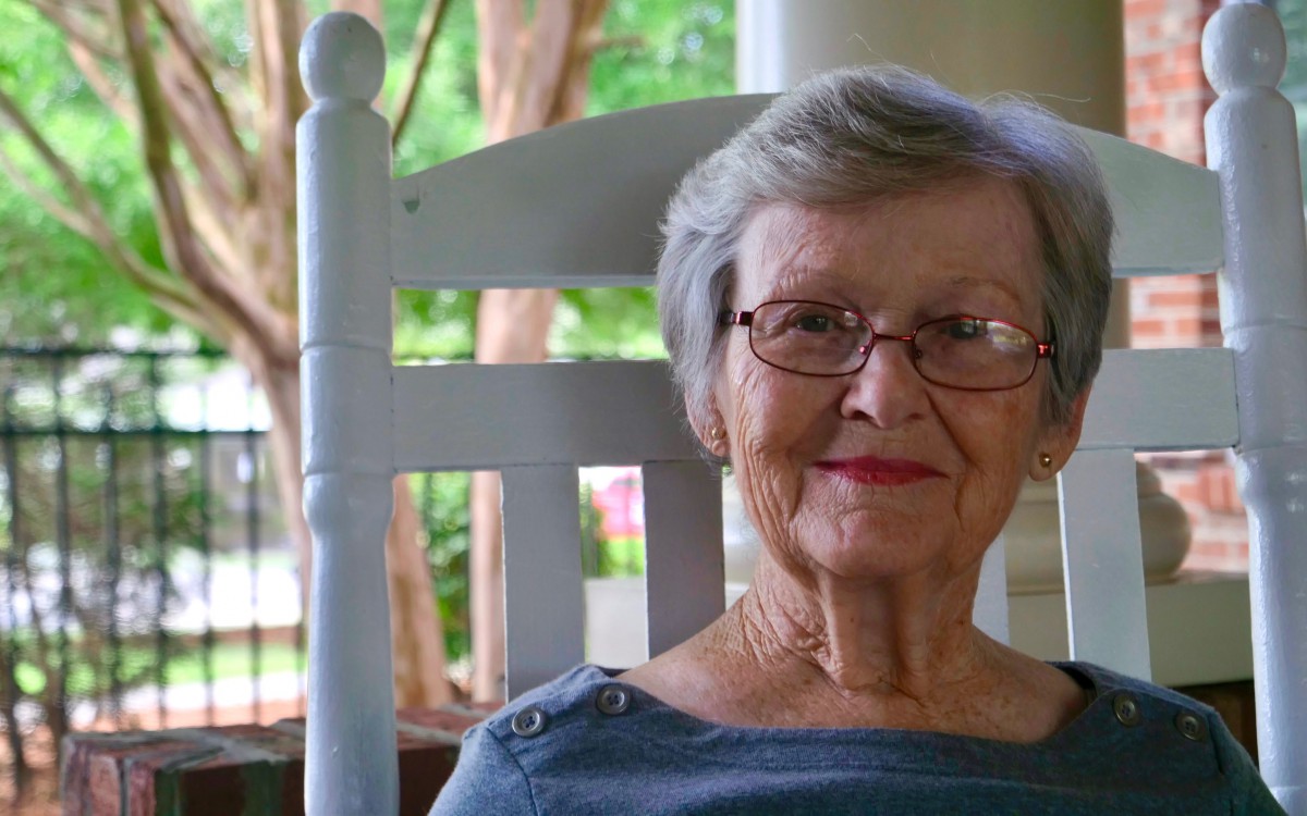 An elderly woman wearing glasses sits comfortably on a porch rocking chair, smiling warmly.