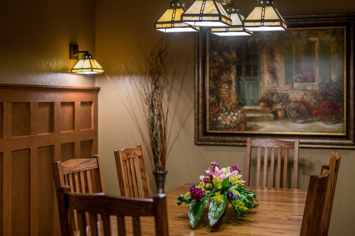 A cozy dining room with wooden furniture, illuminated by a chandelier, features a colorful flower arrangement and a scenic painting on the wall.