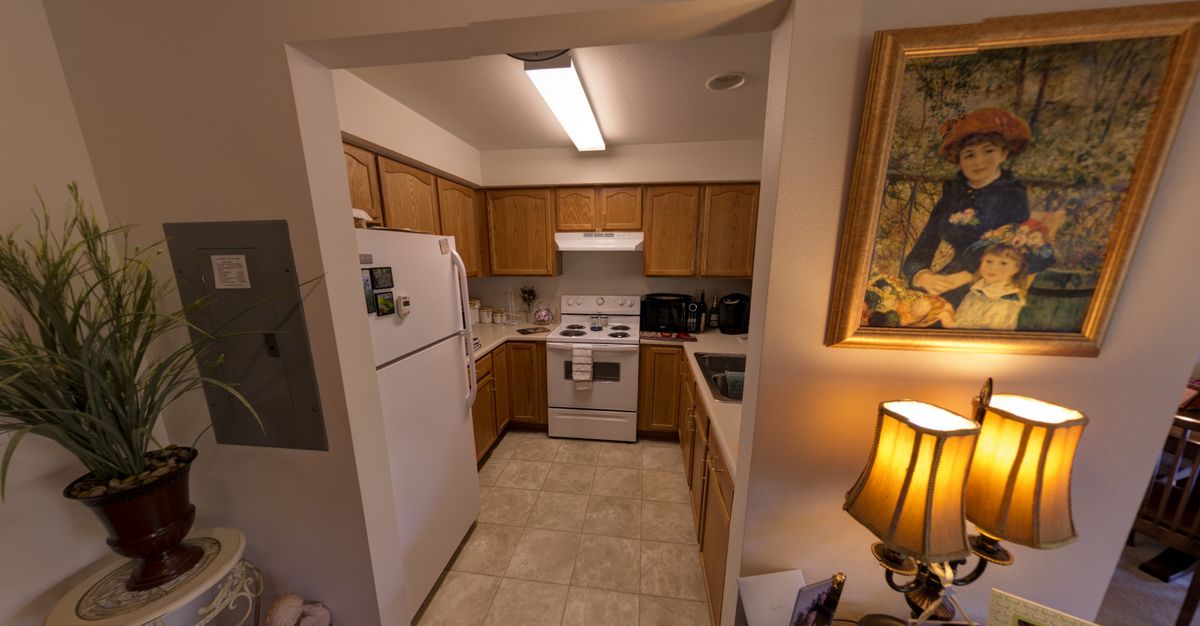 A compact kitchen with wooden cabinets, featuring a refrigerator and stove, illuminated by a ceiling light and accompanied by a lamp and artwork on the wall.