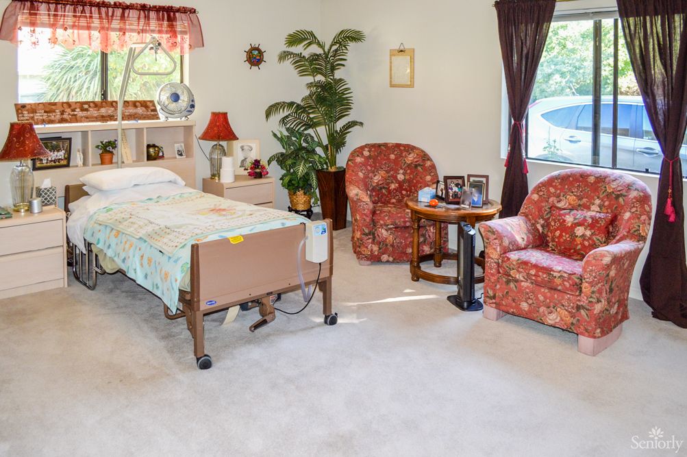 A cozy bedroom with a hospital bed and comfortable seating area, featuring two armchairs and a small table near a large window.