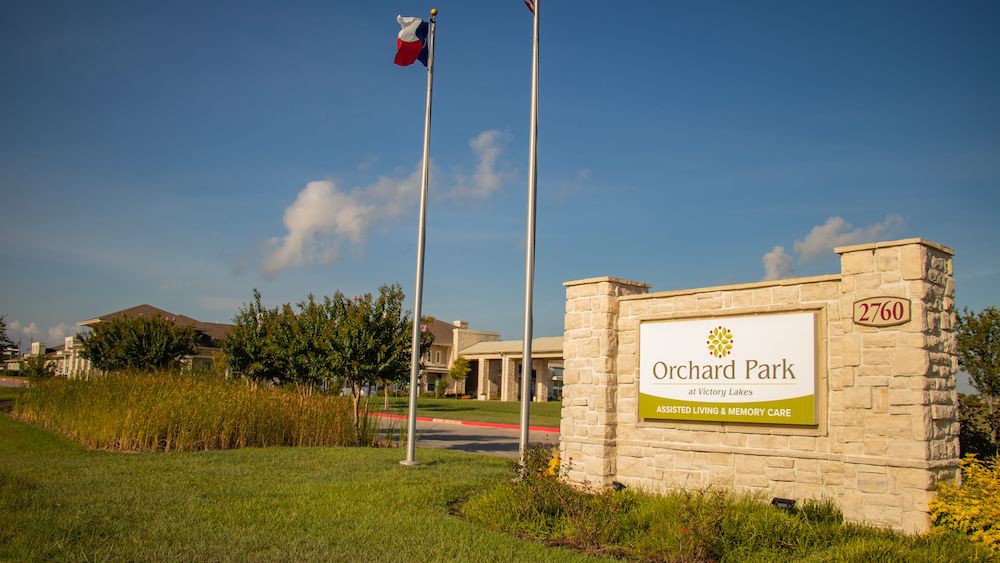 The entrance to Orchard Park, featuring a prominent sign and flagpoles against a blue sky.