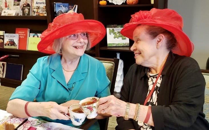 Two elderly women enjoying a tea party, wearing vibrant red hats and sharing smiles.