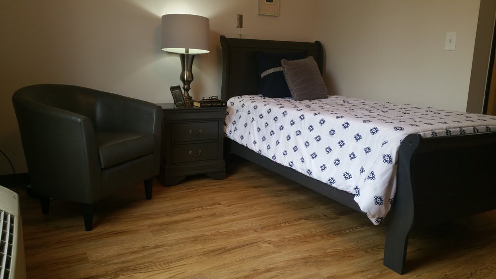 A cozy bedroom featuring a bed with patterned bedding, an armchair, a nightstand with a lamp, and hardwood flooring.